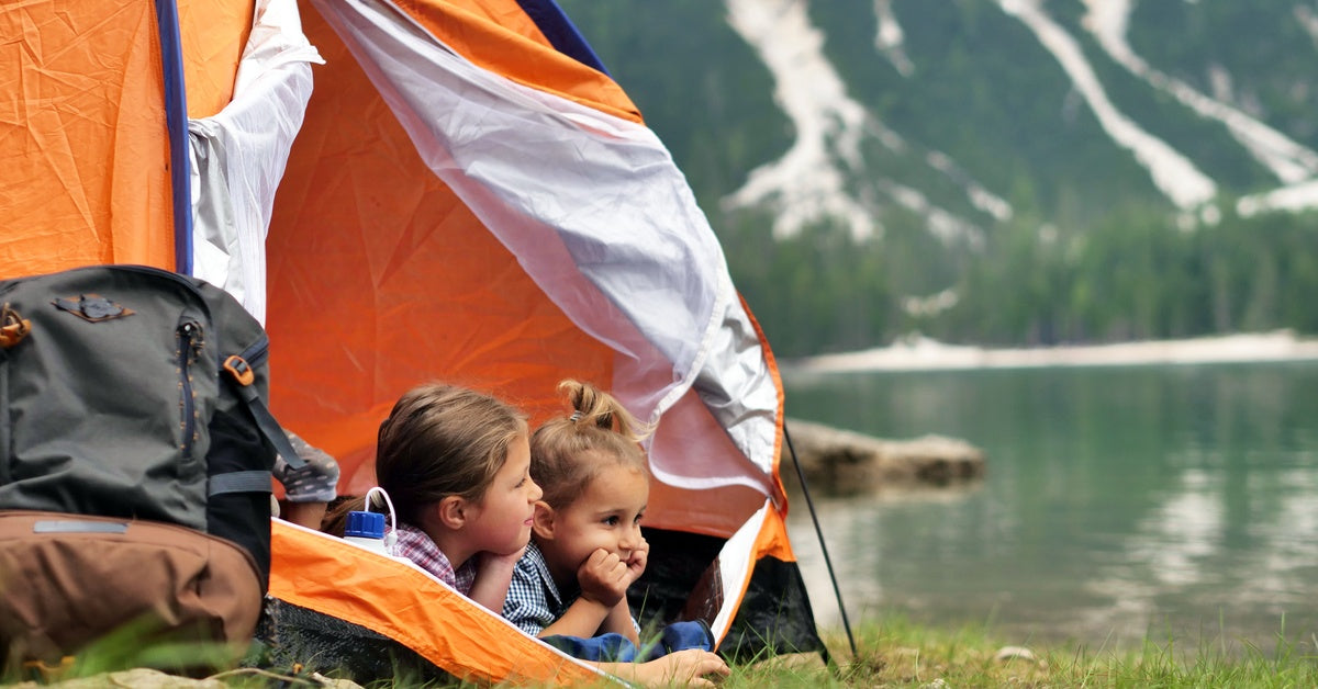 Two little girls lying down in an orange tent, looking out over the landscape. Behind them is a body of water and a mountain.