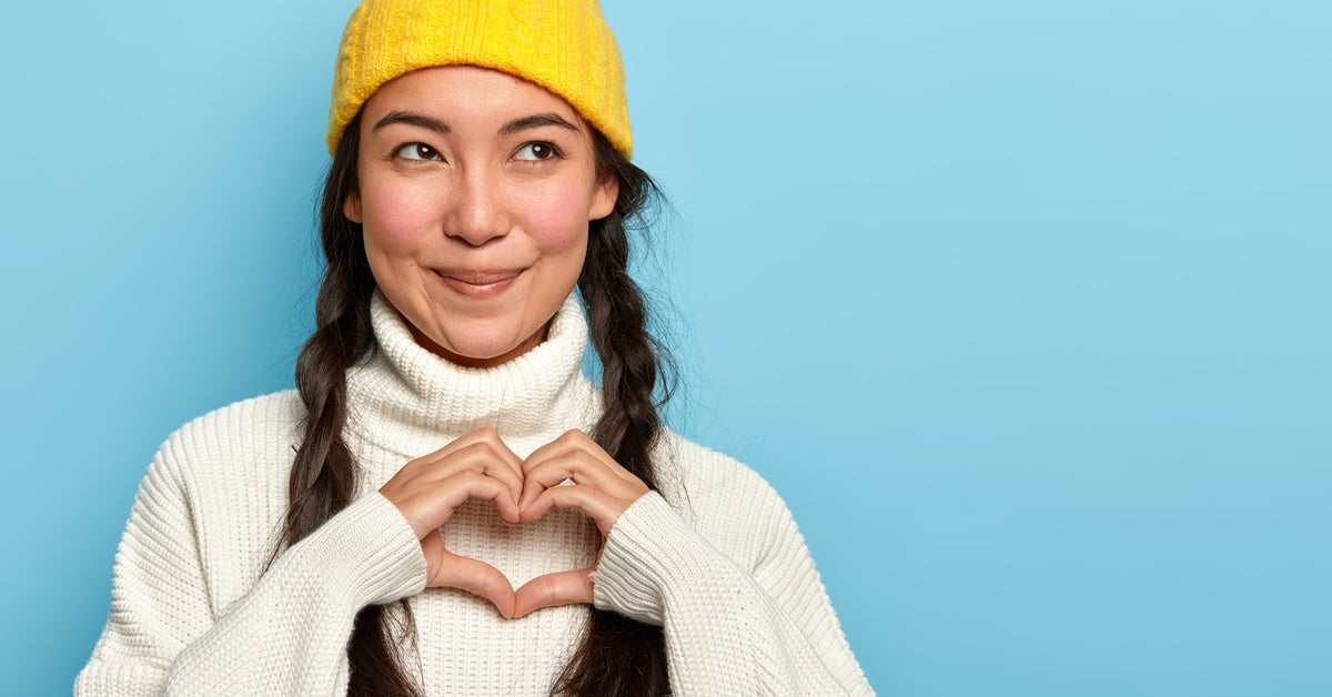 A woman in a yellow beanie with her hair in braids smiling as she makes a heart shape with her hands.