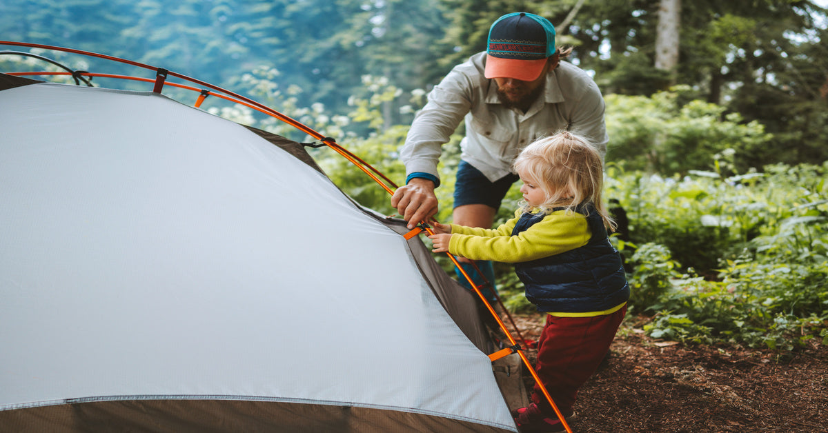 A man and a young child dressed in outdoor wear working together to pitch a tent in the middle of a wooded area.