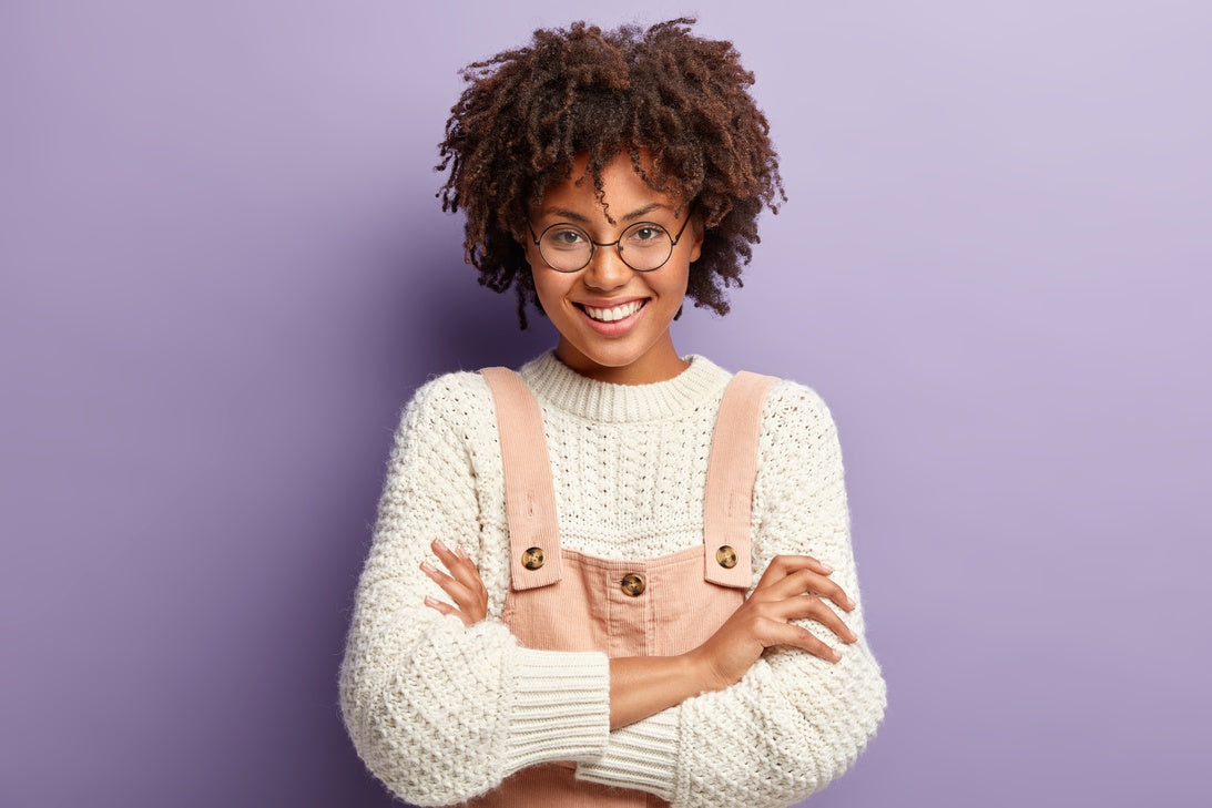 A woman in glasses, a white knit sweater, and dusty pink overalls standing with her arms crossed, posing and smiling.