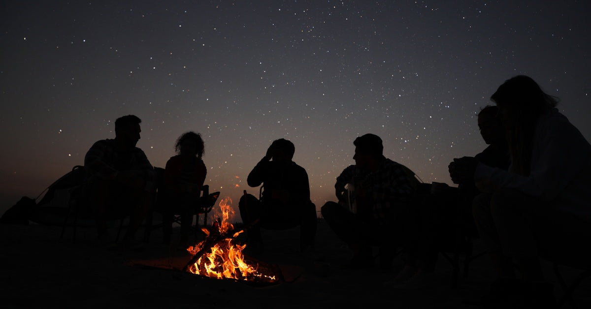 The silhouettes of a group of people sitting around a campfire beneath a beautifully starry night sky.
